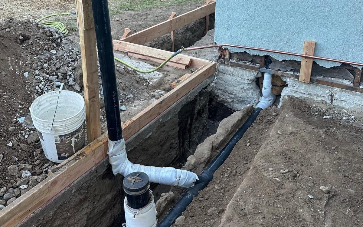A trench along a stucco house wall in a backyard, with new black drain pipe, a white-wrapped cleanout fitting and a tall vent pipe