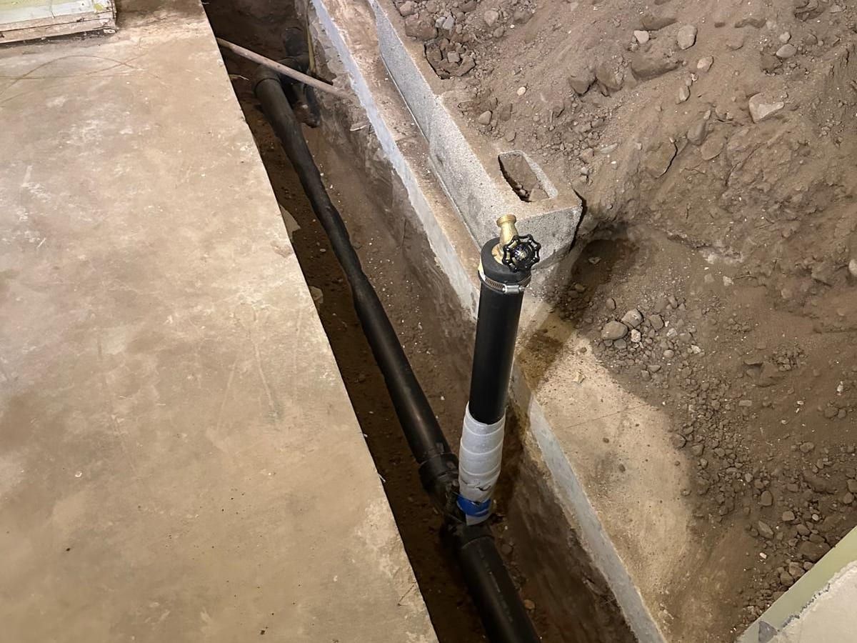 A trench along an inside foundation wall with new black drain pipe and a vertical riser topped with a valve