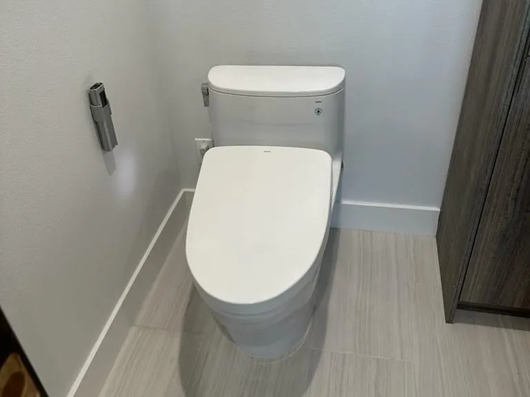 A new white toilet with an elongated seat in a bathroom with white walls and a gray plank tile floor