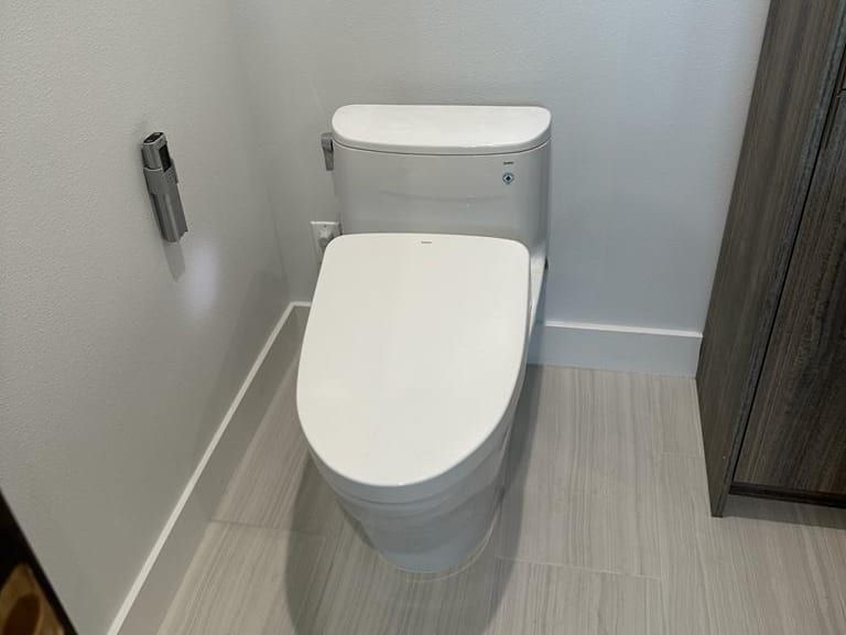 A new white toilet with an elongated seat in a bathroom with white walls and a gray plank tile floor