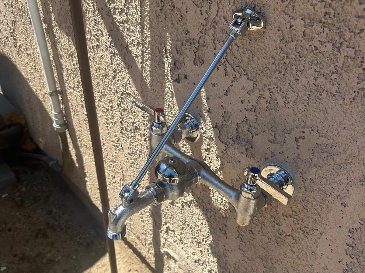 A wall-mounted chrome service faucet with two lever handles, a support rod and a threaded spout, on an exterior stucco wall