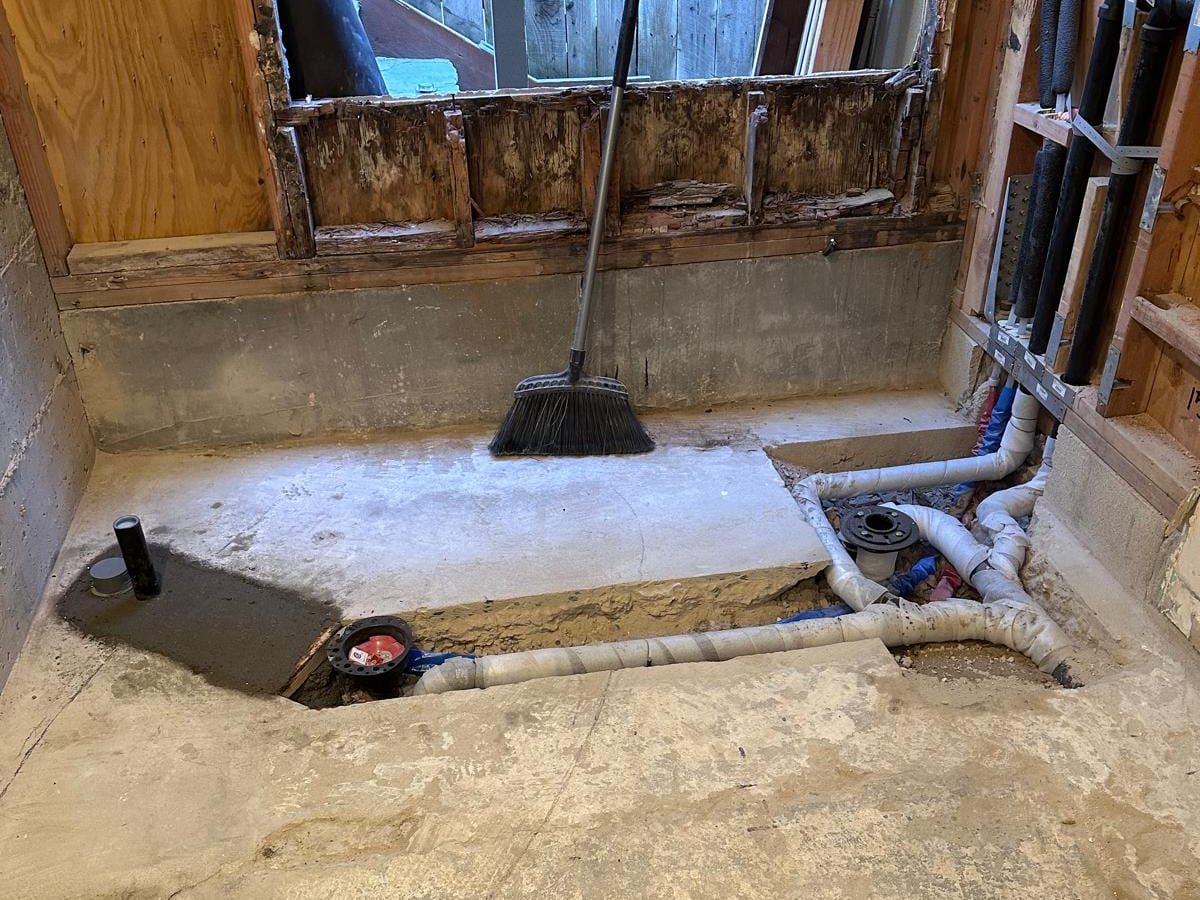 A gutted bathroom with a section of the concrete slab cut open to show new drain lines and a capped toilet flange
