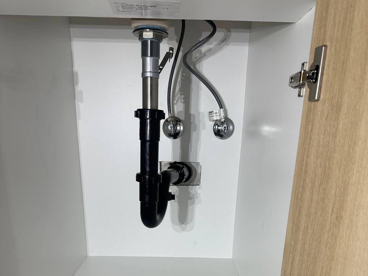 Under a bathroom vanity, a black drain trap, a chrome tailpiece and two supply valves inside a white cabinet